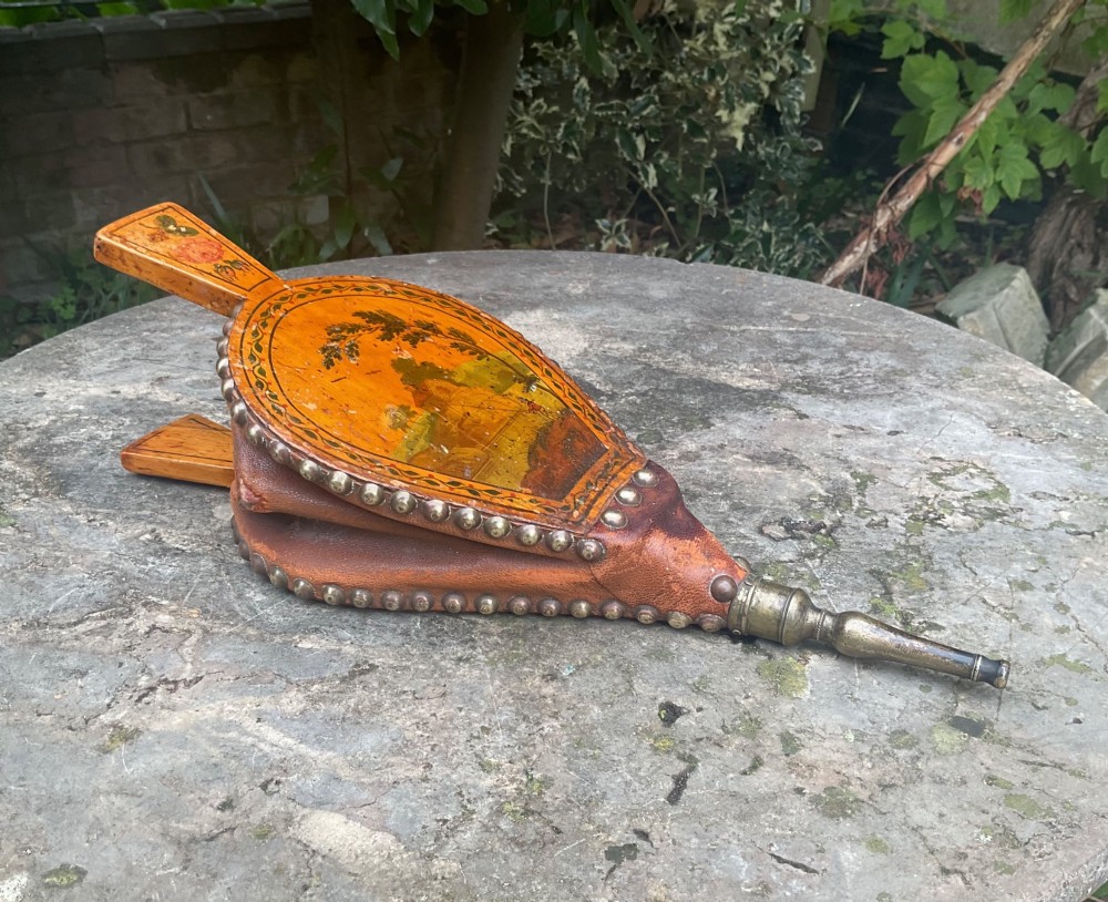 A Regency-period hand-painted wooden fire bellows featuring a painted landscape scene, brass nozzle, tan leather sides, and decorative brass studs.