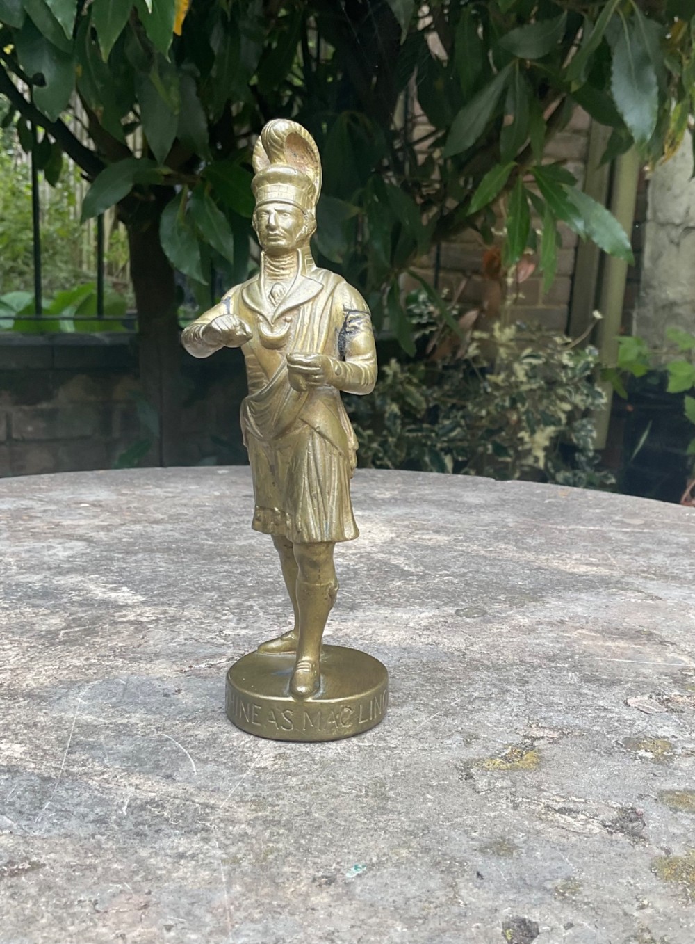 A cast brass car mascot depicting a standing Scottish highlander figure on a circular base inscribed with Phineas MacLino.