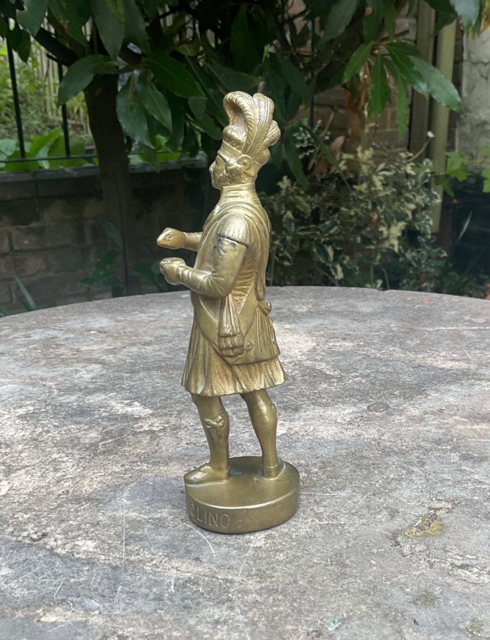 A cast brass car mascot depicting a standing Scottish highlander figure on a circular base inscribed with Phineas MacLino. View II
