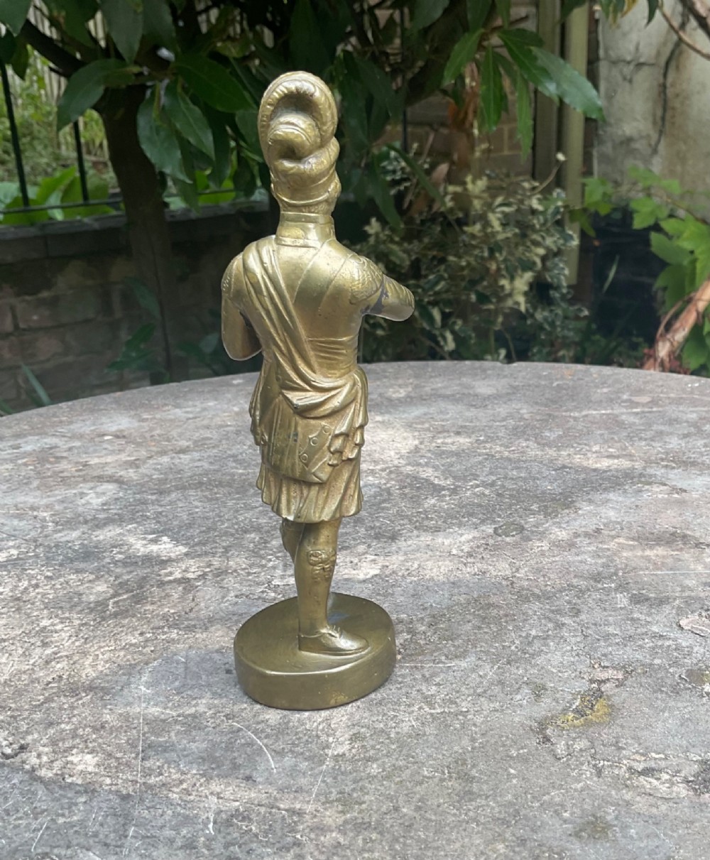 A cast brass car mascot depicting a standing Scottish highlander figure on a circular base inscribed with Phineas MacLino. View III