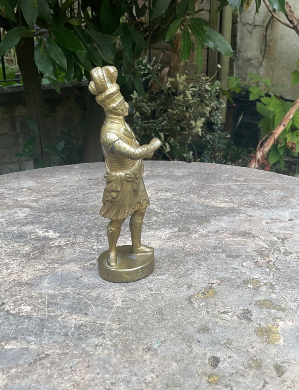 A cast brass car mascot depicting a standing Scottish highlander figure on a circular base inscribed with Phineas MacLino. View IV