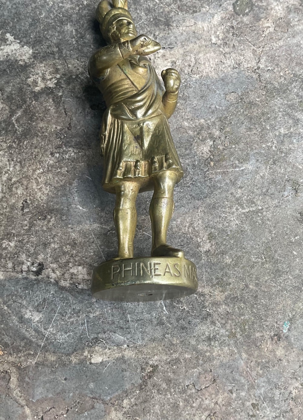 A cast brass car mascot depicting a standing Scottish highlander figure on a circular base inscribed with Phineas MacLino. View VI