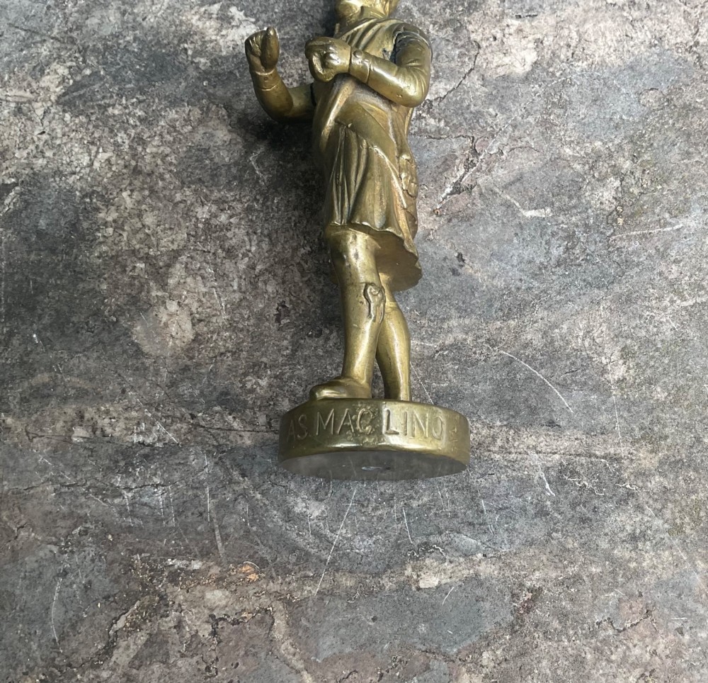 A cast brass car mascot depicting a standing Scottish highlander figure on a circular base inscribed with Phineas MacLino. View VII