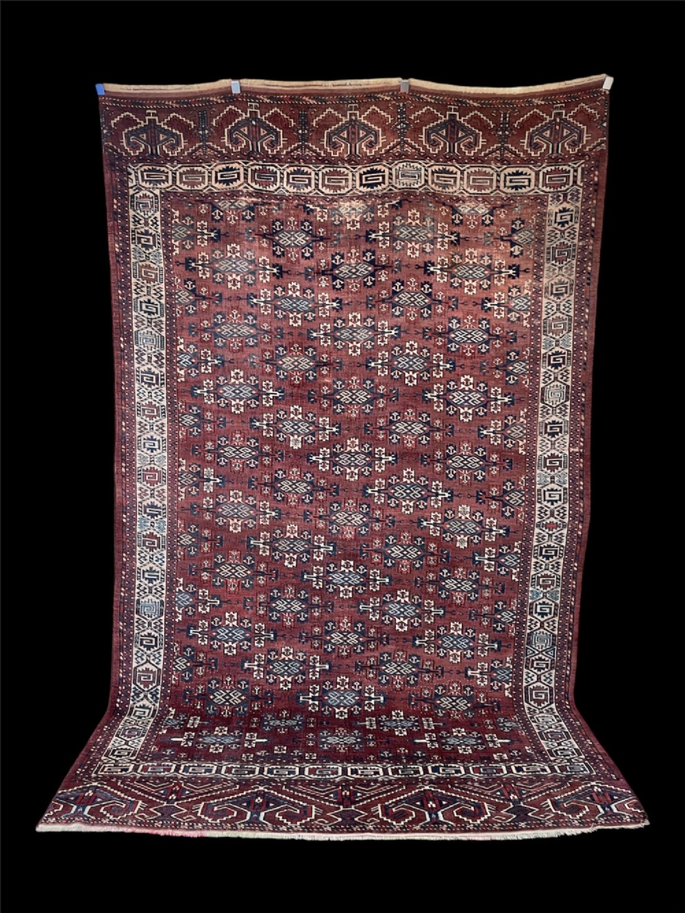 A large antique hand-knotted wool Turkoman Yomut carpet featuring repeating geometric gol motifs on a deep red ground surrounded by geometric borders.
