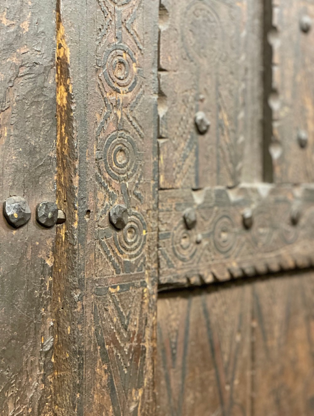 A hand-carved dark timber Amazigh door featuring geometric relief patterns, wrought iron studs, and a circular iron knocker. View IV