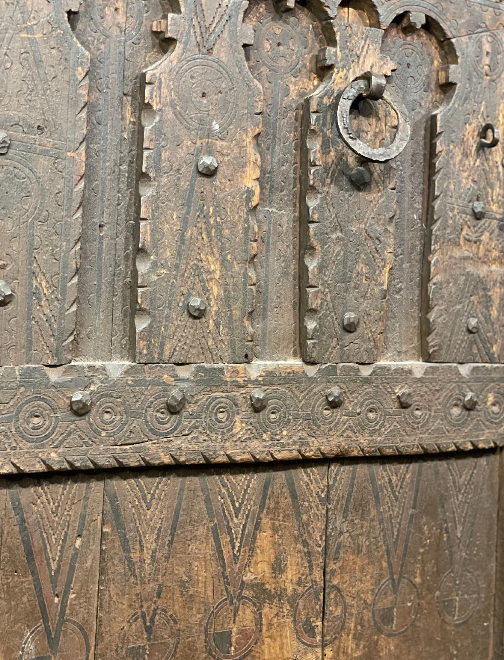 A hand-carved dark timber Amazigh door featuring geometric relief patterns, wrought iron studs, and a circular iron knocker. View V