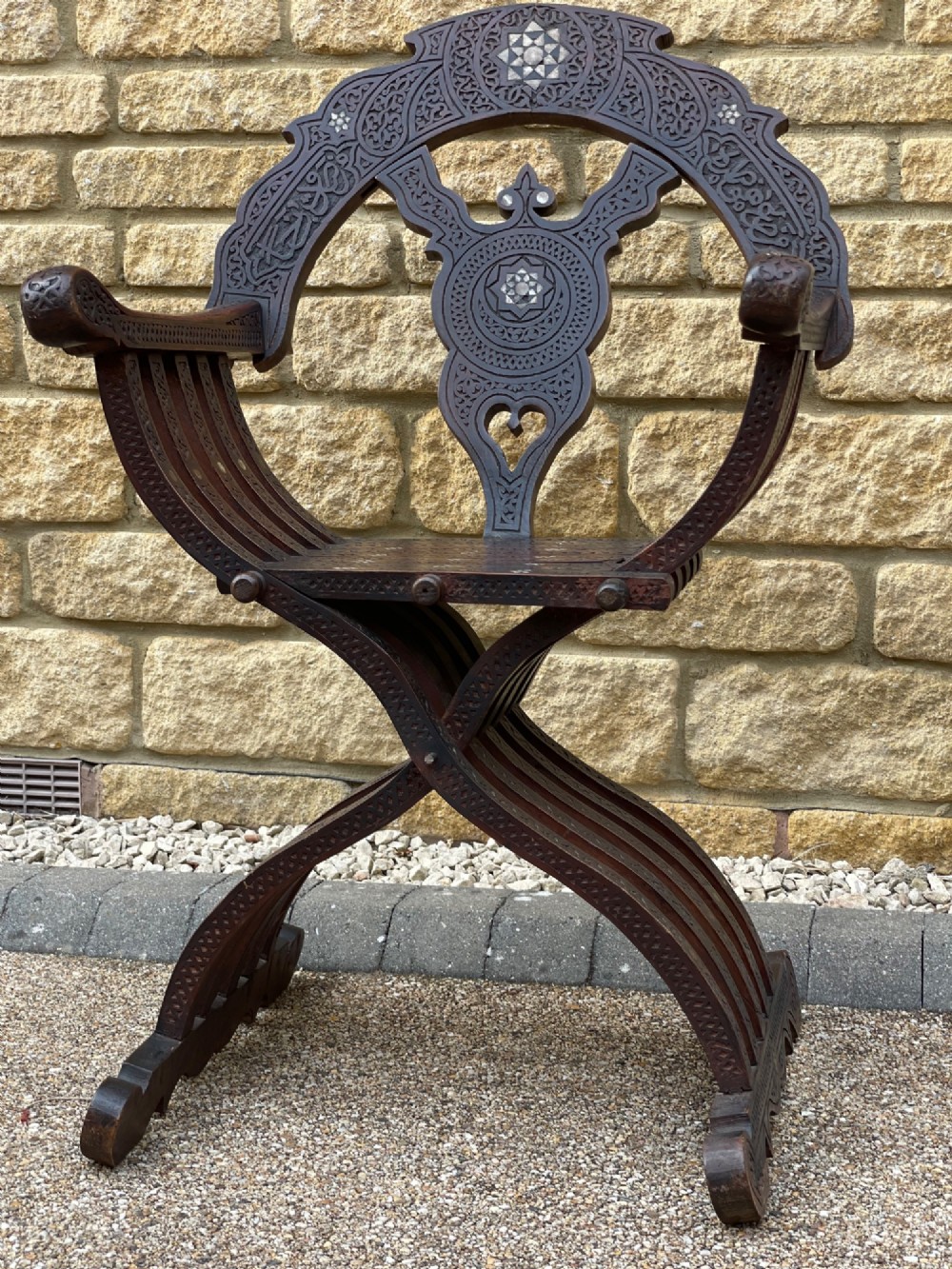 A dark carved wood X-frame armchair featuring intricate Arabic geometric fretwork and inlaid star motifs crafted from iridescent mother of pearl.