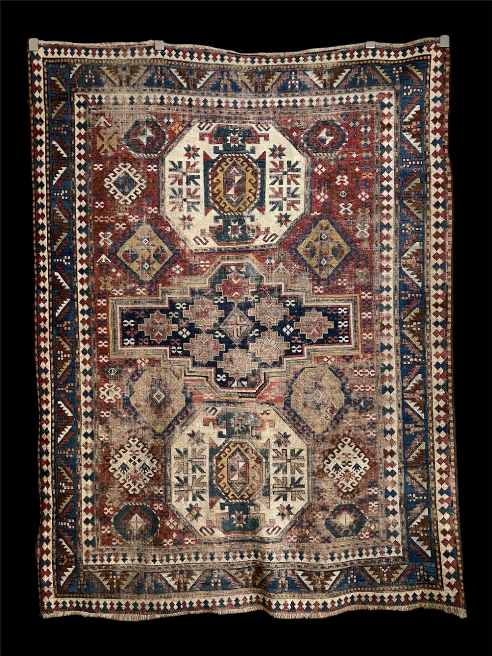 An antique Caucasian Kazak hand-knotted wool rug featuring geometric stepped medallions and latch-hook borders in shades of red, indigo blue, and cream.
