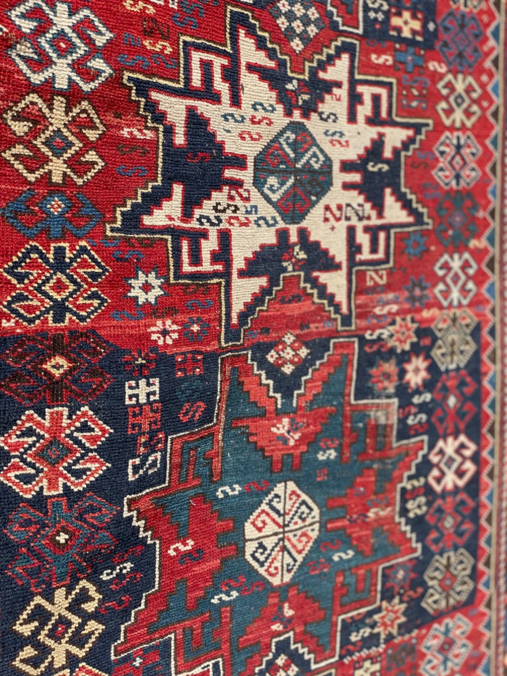 An antique Caucasian hand-knotted wool rug featuring geometric Lezghi star medallions in red, indigo blue, and ivory framed by patterned geometric borders. View III