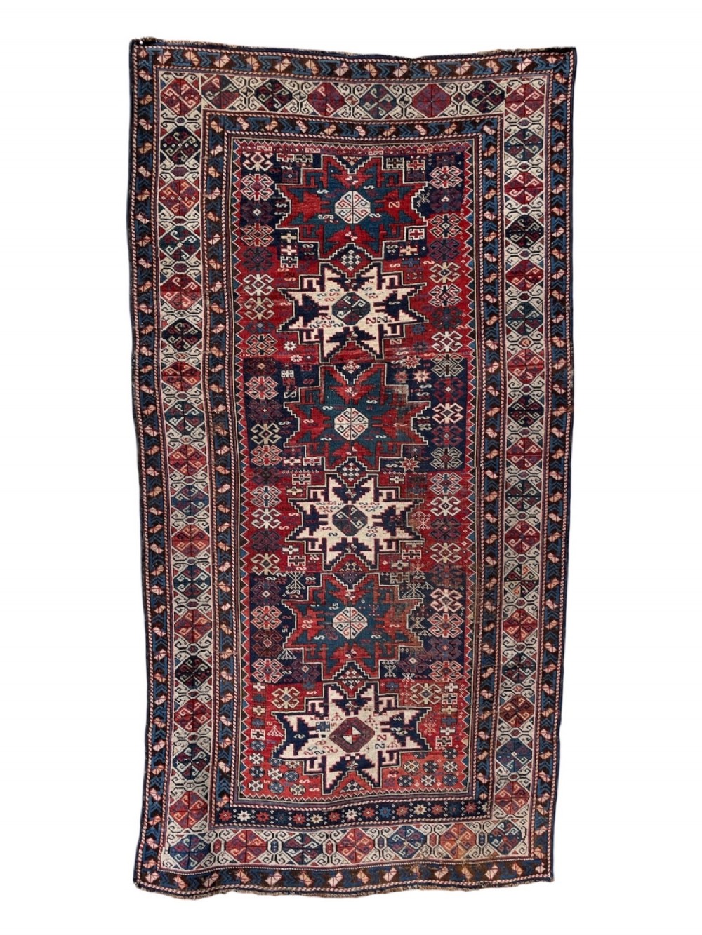 An antique Caucasian hand-knotted wool rug featuring geometric Lezghi star medallions in red, indigo blue, and ivory framed by patterned geometric borders.