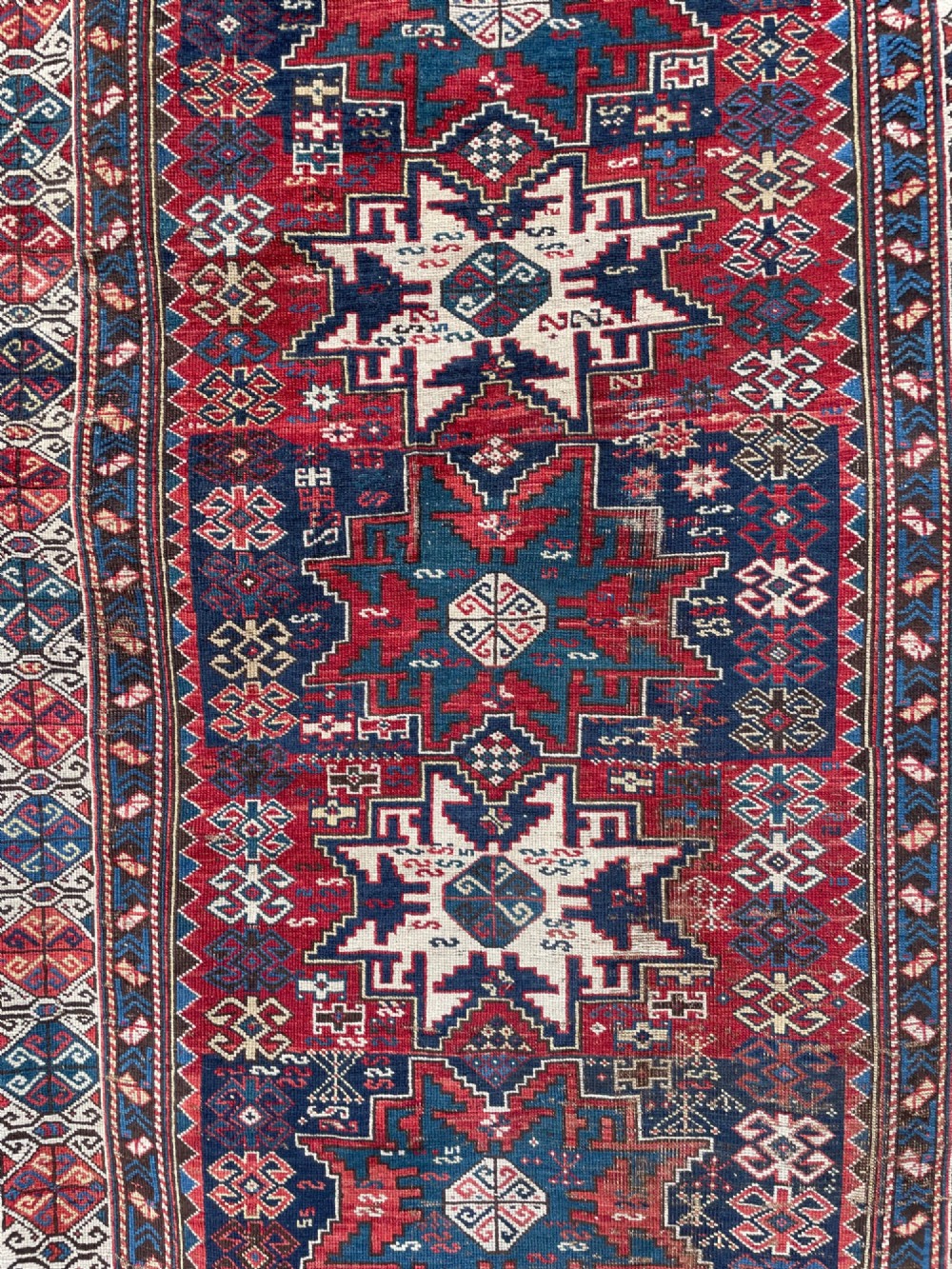 An antique Caucasian hand-knotted wool rug featuring geometric Lezghi star medallions in red, indigo blue, and ivory framed by patterned geometric borders. View IV