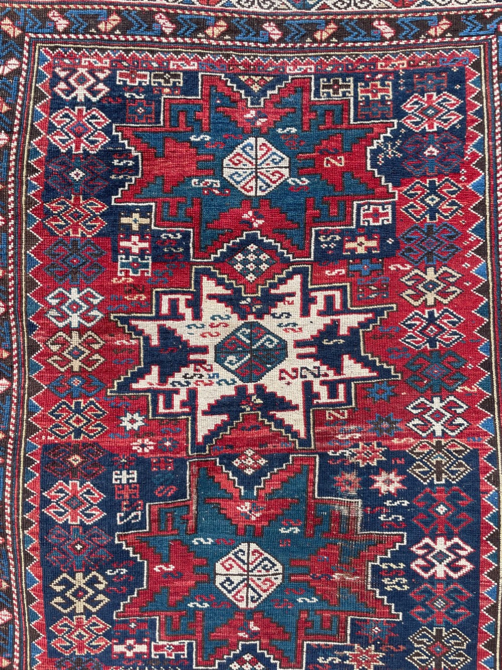 An antique Caucasian hand-knotted wool rug featuring geometric Lezghi star medallions in red, indigo blue, and ivory framed by patterned geometric borders. View V