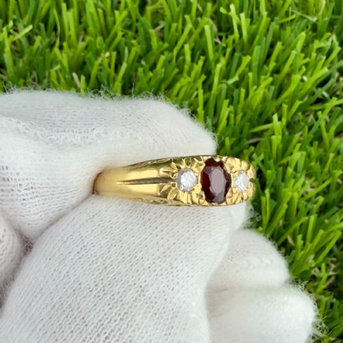 fig 1. An 18ct yellow gold trilogy ring featuring a central oval dark red garnet flanked by two round diamonds in carved star settings on a wide band.