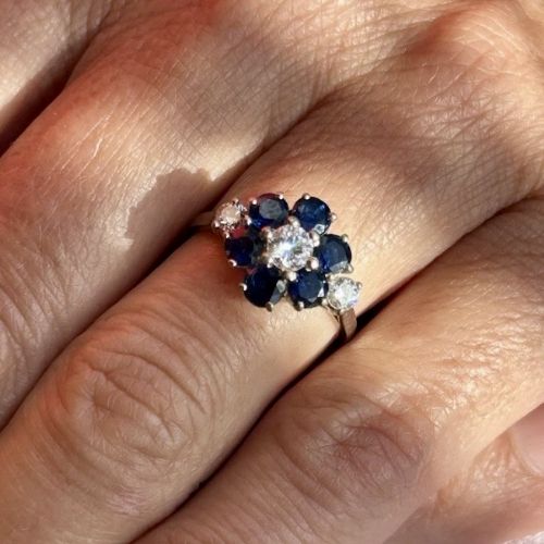 fig 1. An 18ct white gold cluster ring featuring a central round diamond surrounded by six round dark blue sapphires in a flower motif with diamond accents on the shoulders.
