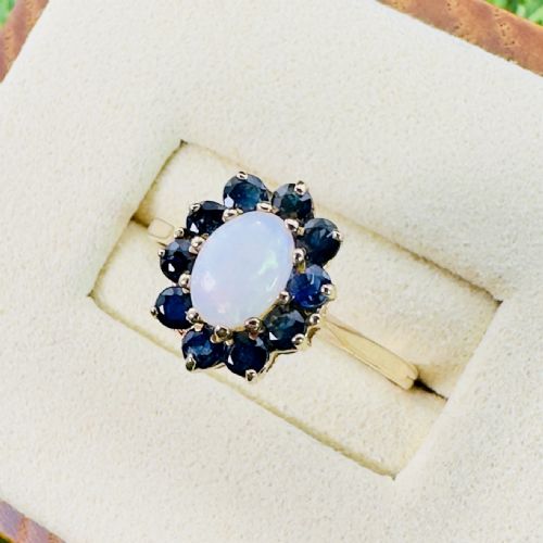 fig 1. A 9ct yellow gold ring featuring a central oval cabochon opal surrounded by ten round blue sapphires in claw settings.