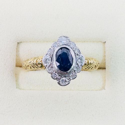 fig 1. An 18ct yellow gold ring featuring an oval cut deep blue sapphire framed by a halo of diamonds on an intricately engraved band.