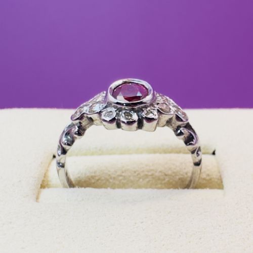 fig 1. An 18ct white gold cluster ring featuring a central oval red ruby surrounded by small round diamonds and carved shoulder details.