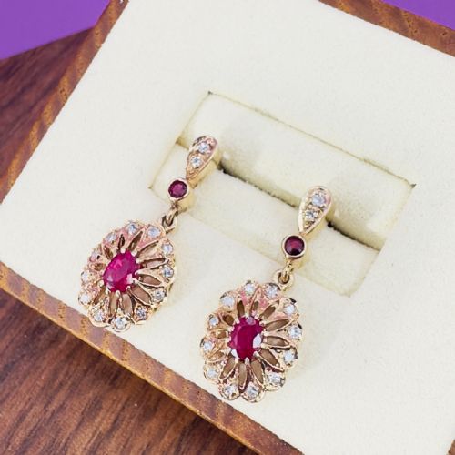 fig 1. A pair of 9ct gold drop stud earrings featuring central oval red rubies surrounded by diamond clusters in a sunburst design.