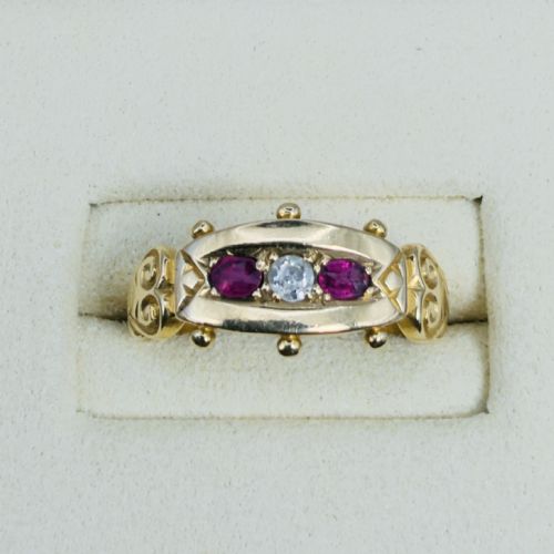 fig 1. An 18ct yellow gold Edwardian trilogy ring set with a central round diamond flanked by two oval red rubies, featuring scroll detailing on the shoulders.