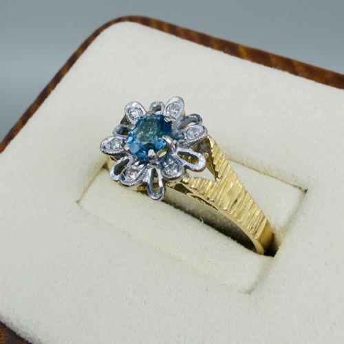 fig 1. An 18ct yellow gold ring featuring a central oval blue aquamarine framed by an openwork floral halo set with diamonds and textured shoulders.