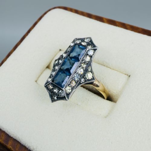 fig 1. An Art Deco 18ct gold ring featuring a central vertical row of three square blue stones framed by white paste stones in an elongated geometric silver setting.