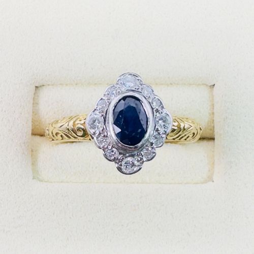 fig 1. An 18ct yellow gold ring featuring a central oval blue sapphire surrounded by a border of small diamonds, set on a carved scrollwork band.