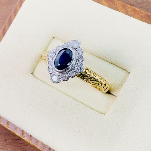 fig 2. An 18ct yellow gold ring featuring a central oval blue sapphire surrounded by a border of small diamonds, set on a carved scrollwork band.