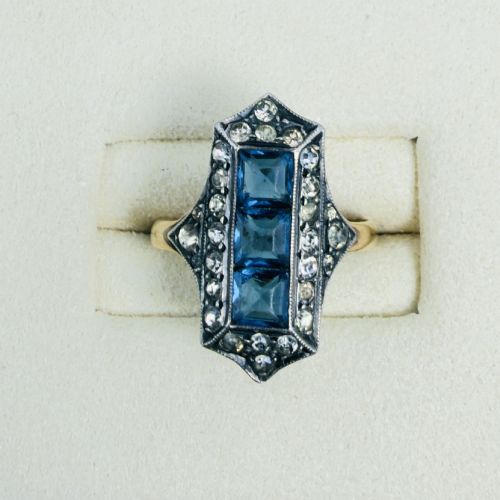 fig 1. An Art Deco dress ring featuring an elongated geometric silver top set with three rectangular blue stones and bordered by round white paste stones on a yellow gold band.