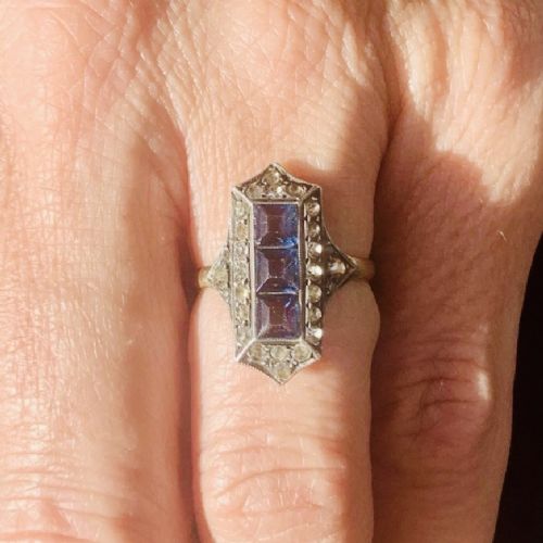 An Art Deco dress ring featuring an elongated geometric silver top set with three rectangular blue stones and bordered by round white paste stones on a yellow gold band. View XII