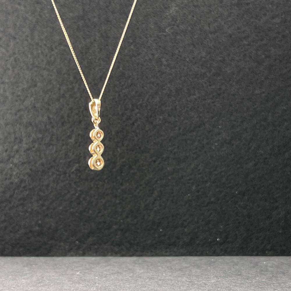 A 9ct yellow gold fine link chain necklace featuring a vertical drop pendant set with three round brilliant cut diamonds in bezel settings. View V