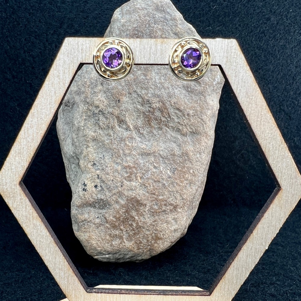 A pair of circular 9ct gold stud earrings set with dark purple amethysts and surrounded by patterned gold borders, alongside gold butterfly backs. View IV
