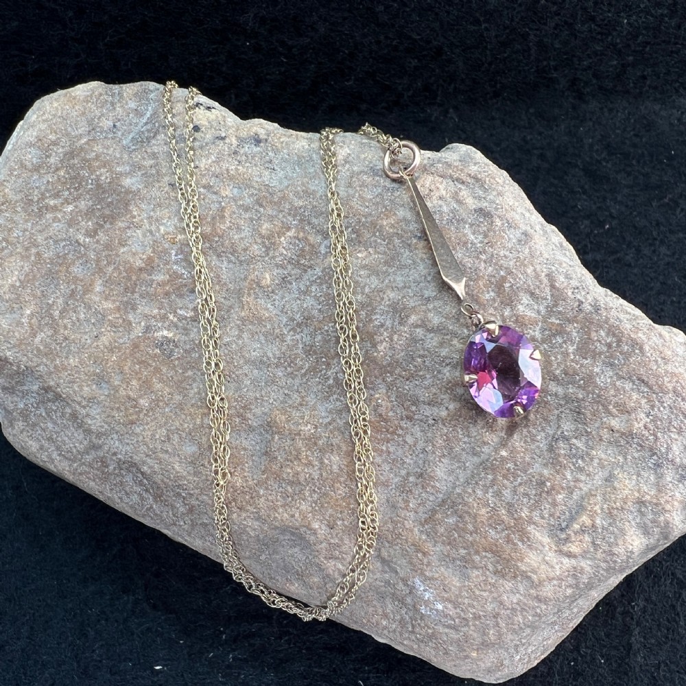 A 9ct gold pendant necklace featuring an elongated tapered bail drop suspending an oval faceted purple amethyst in a claw setting. View XII