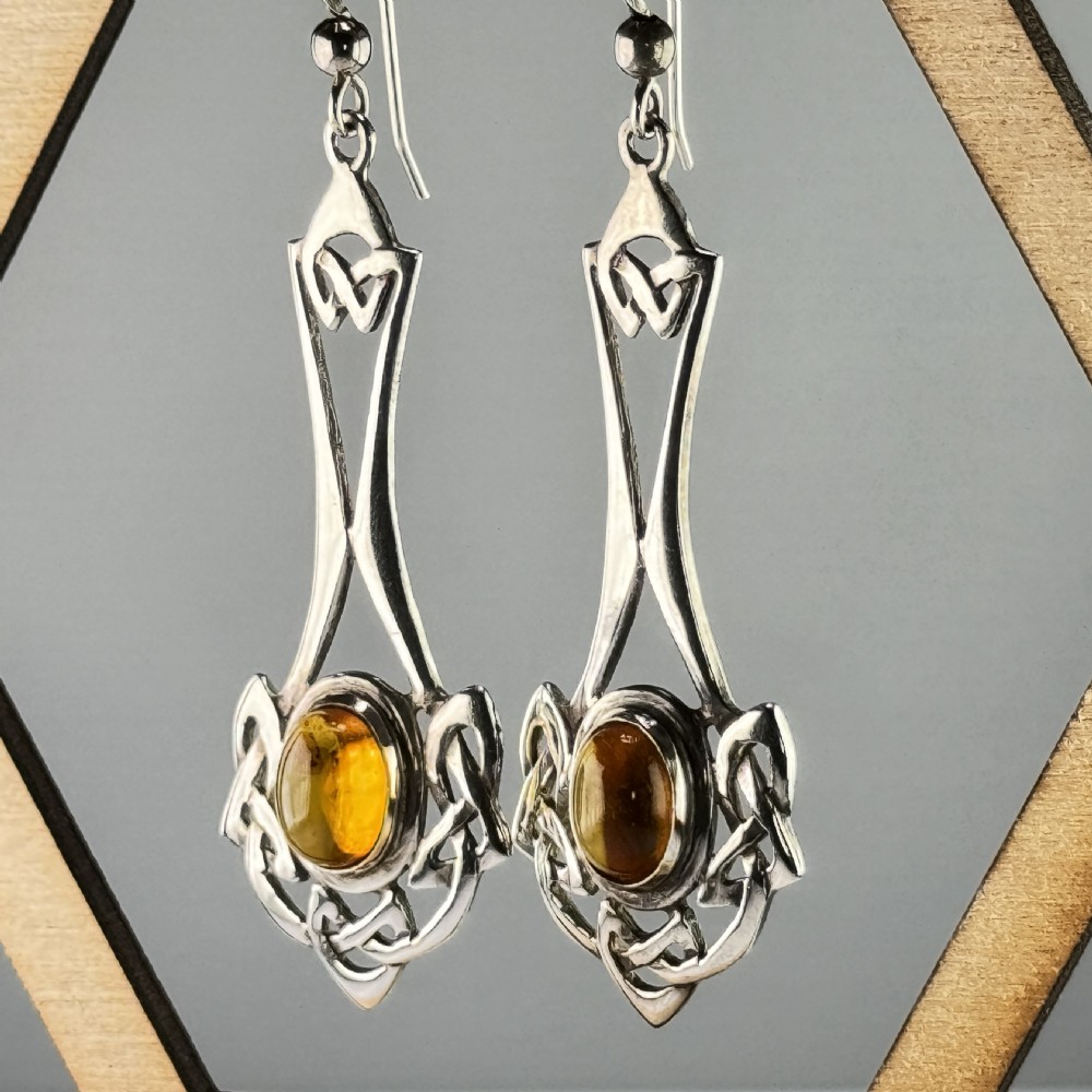 A pair of sterling silver drop earrings featuring intricate openwork Celtic knot patterns encasing oval amber cabochon stones. View II