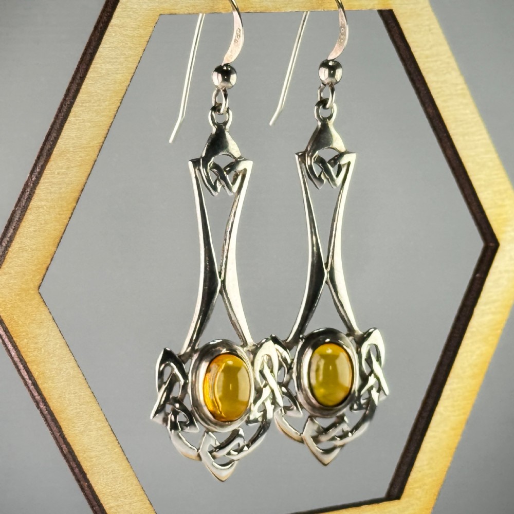 A pair of sterling silver drop earrings featuring intricate openwork Celtic knot patterns encasing oval amber cabochon stones. View V
