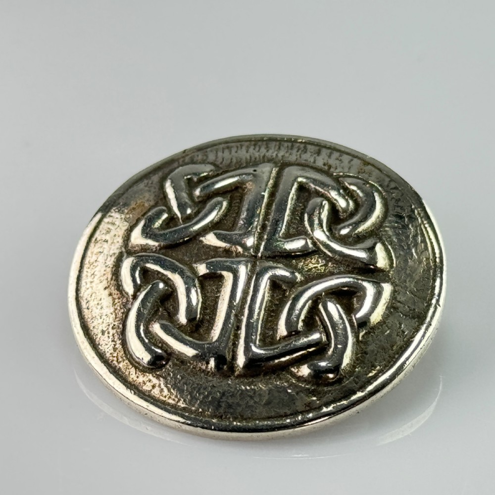 A circular silver brooch featuring raised Celtic knotwork patterns against a textured oxidised background. View V