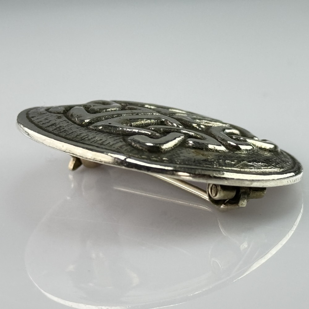 A circular silver brooch featuring raised Celtic knotwork patterns against a textured oxidised background. View VI