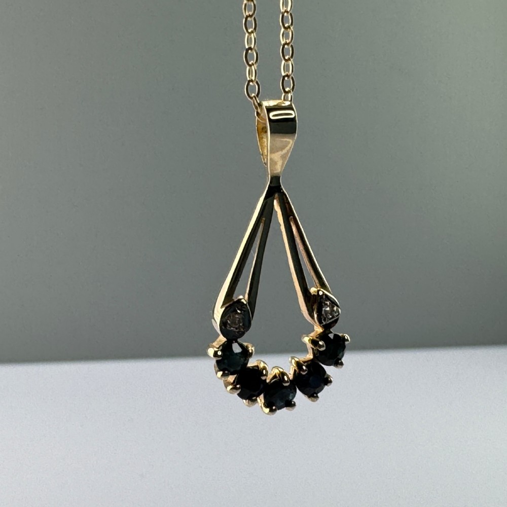 A 9ct yellow gold openwork drop pendant set with five round dark blue sapphires and two small accent diamonds on a fine chain. View V