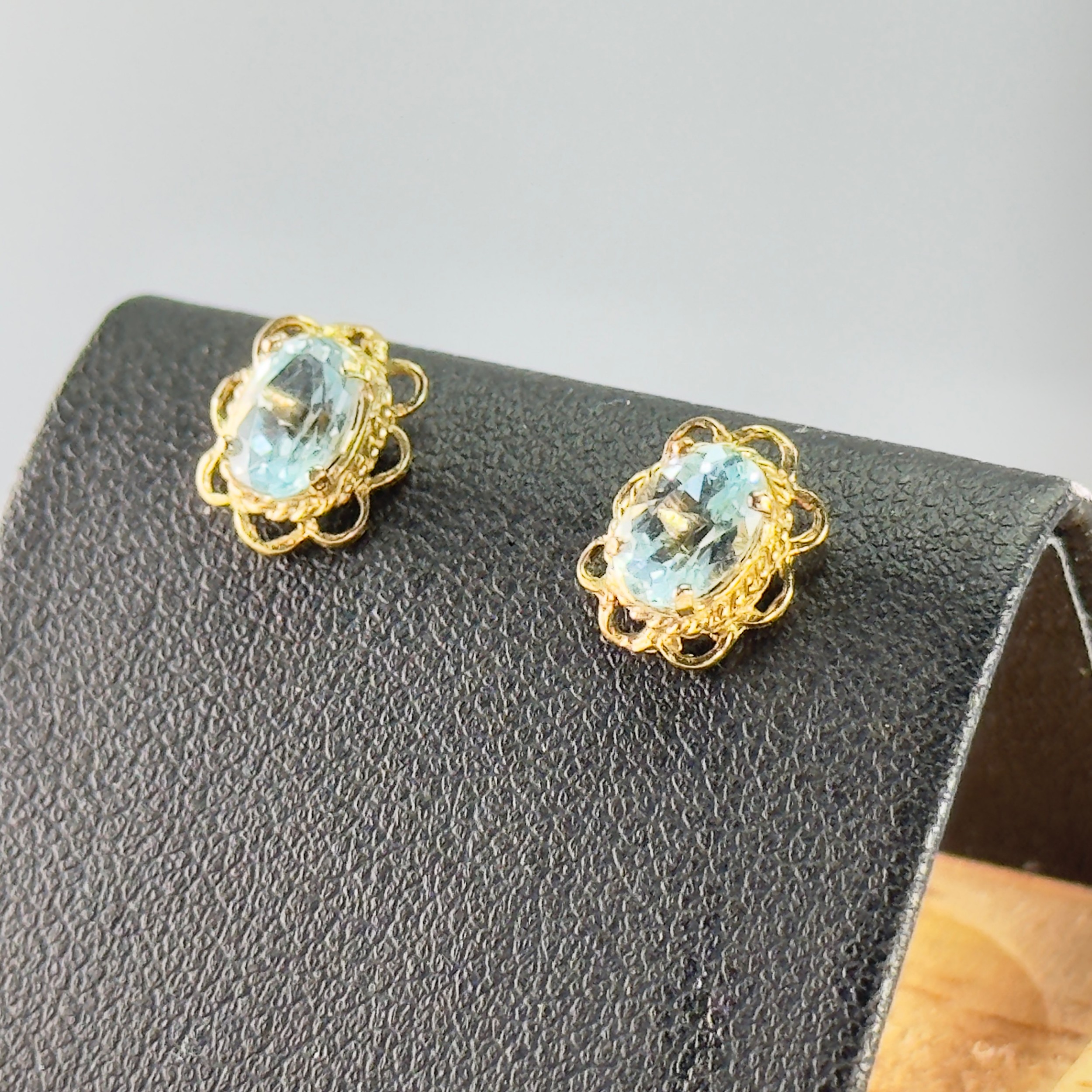 A pair of 9ct yellow gold stud earrings featuring oval light blue topaz gemstones framed by intricate scalloped filigree borders.