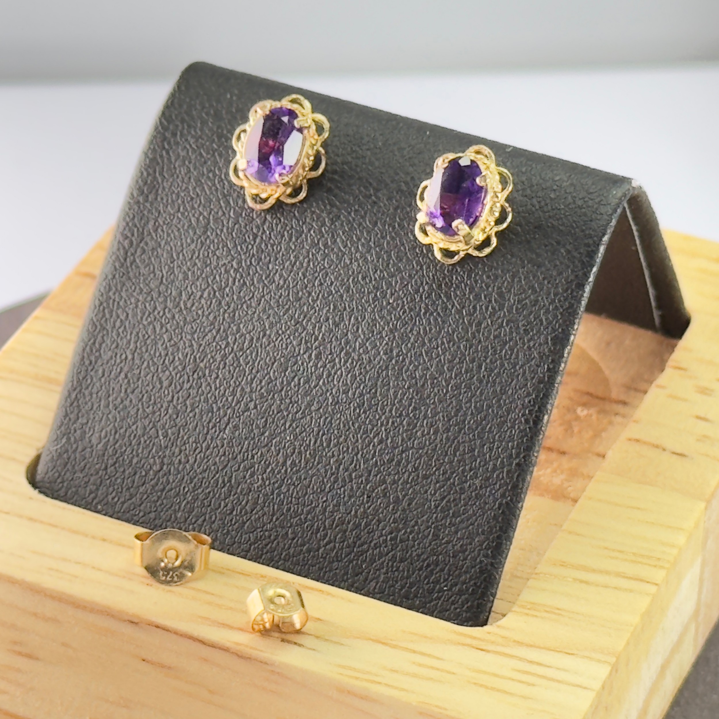 A pair of 9ct yellow gold stud earrings featuring oval cut purple amethyst gemstones surrounded by decorative scalloped filigree borders.