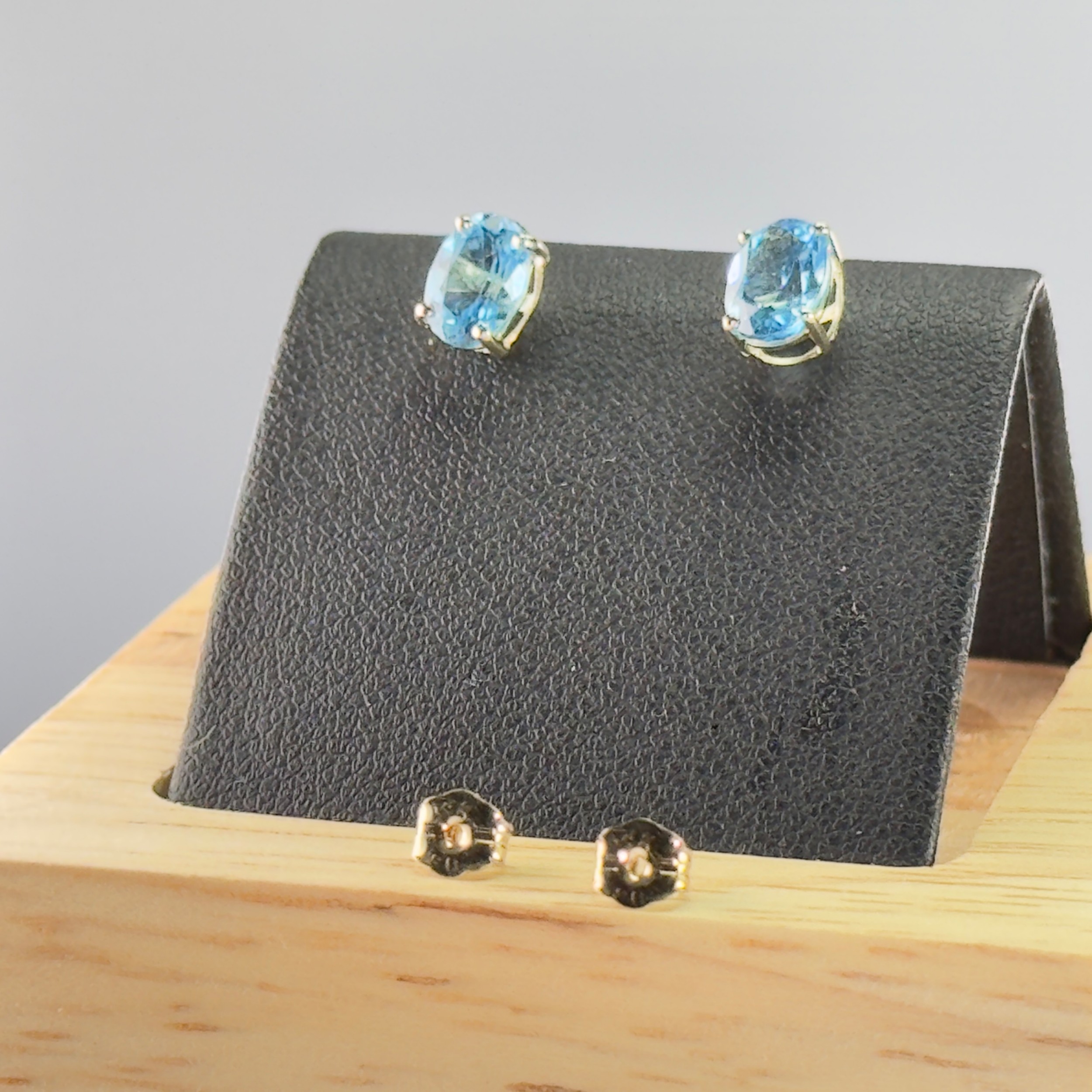 A pair of oval-cut light blue topaz gemstones set in four-prong 9ct yellow gold stud earrings alongside matching butterfly backs.
