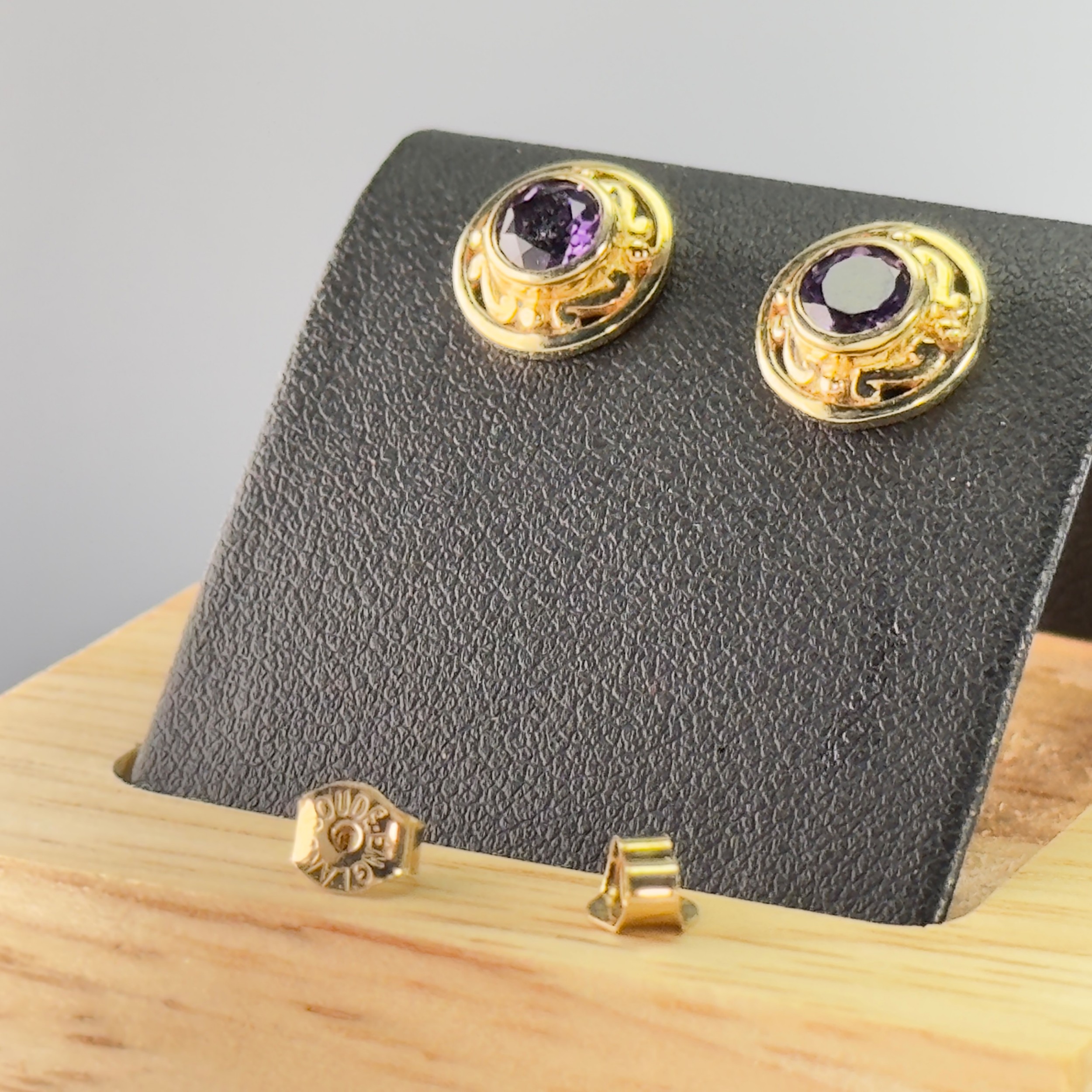 A pair of circular 9ct gold stud earrings set with dark purple amethysts and surrounded by patterned gold borders, alongside gold butterfly backs.