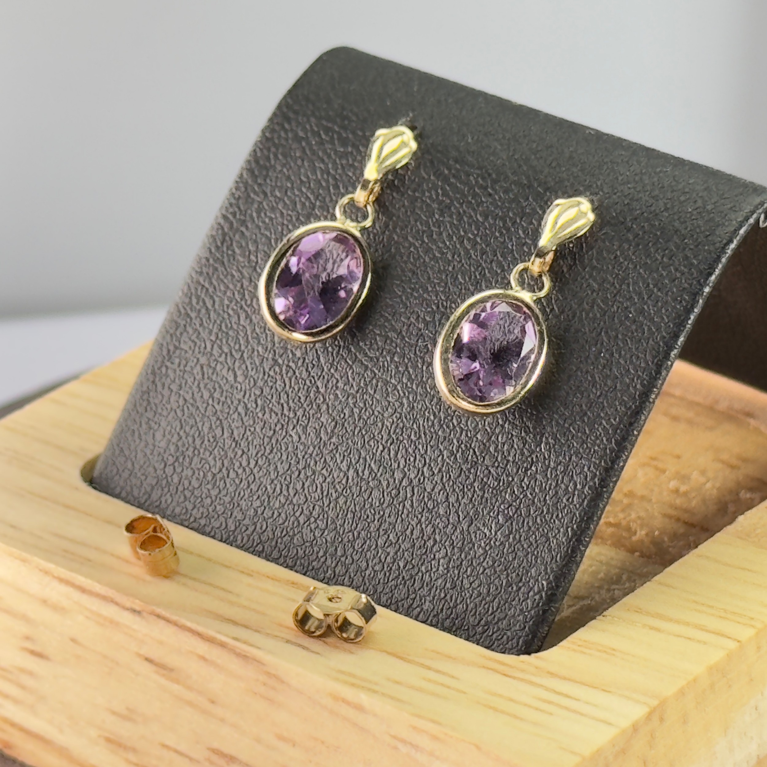 Pair of 9ct yellow gold drop earrings featuring oval bezel-set amethyst gemstones with textured stud tops and scroll backs.