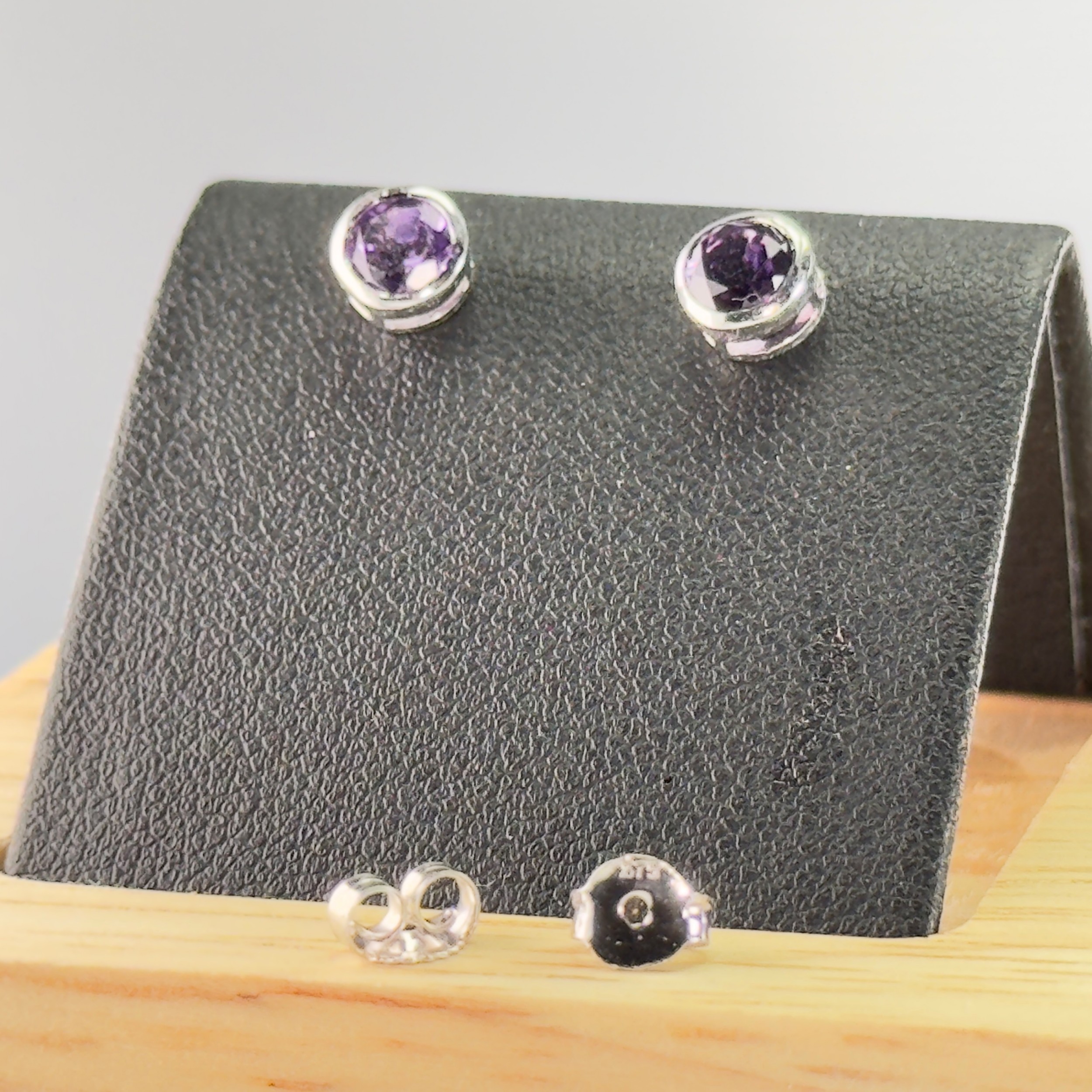 A pair of 9ct white gold stud earrings featuring round bezel-set purple amethyst gemstones accompanied by scroll back fittings.