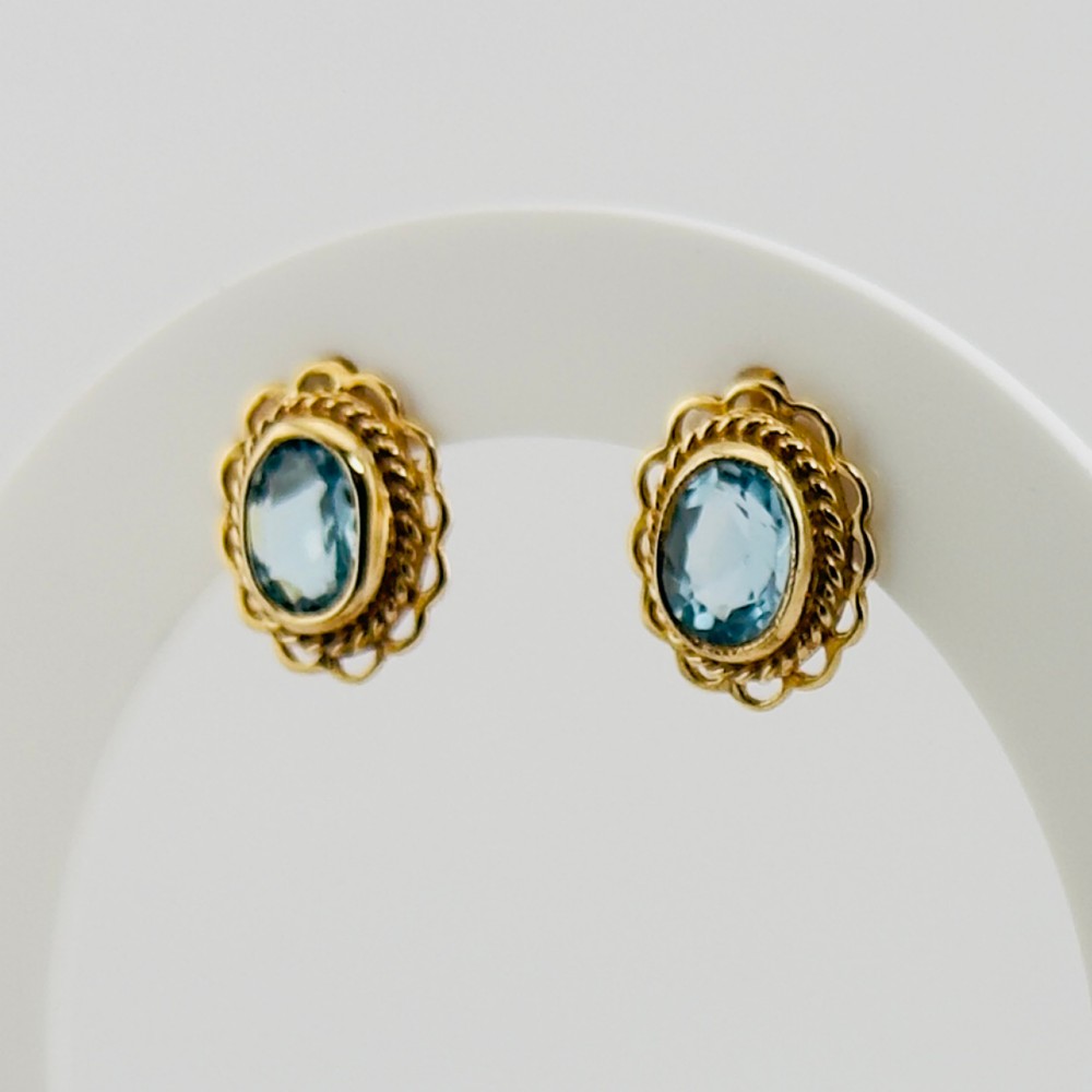A pair of 9ct yellow gold stud earrings featuring oval blue topaz gemstones encased within filigree and rope-twist borders.