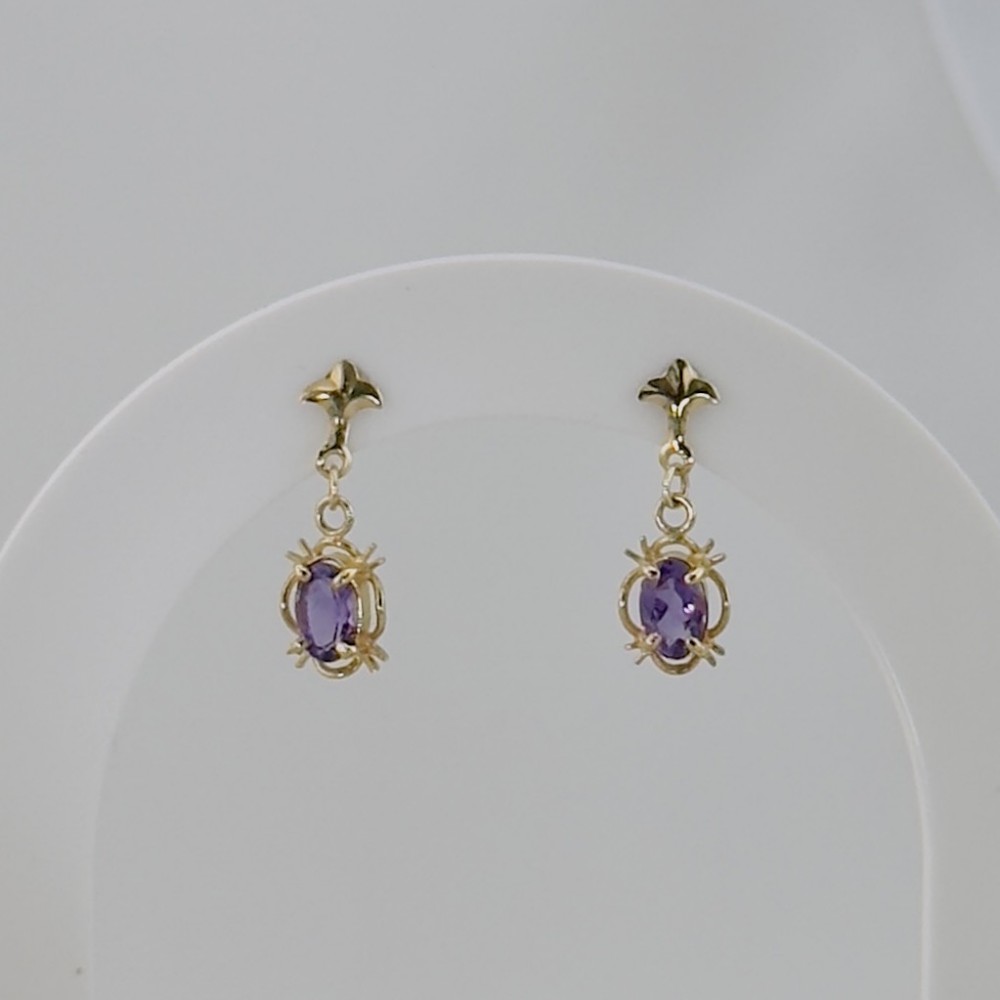 A pair of yellow gold drop earrings, each featuring an oval faceted purple amethyst gemstone set in a clawed frame suspended from a decorative stud.