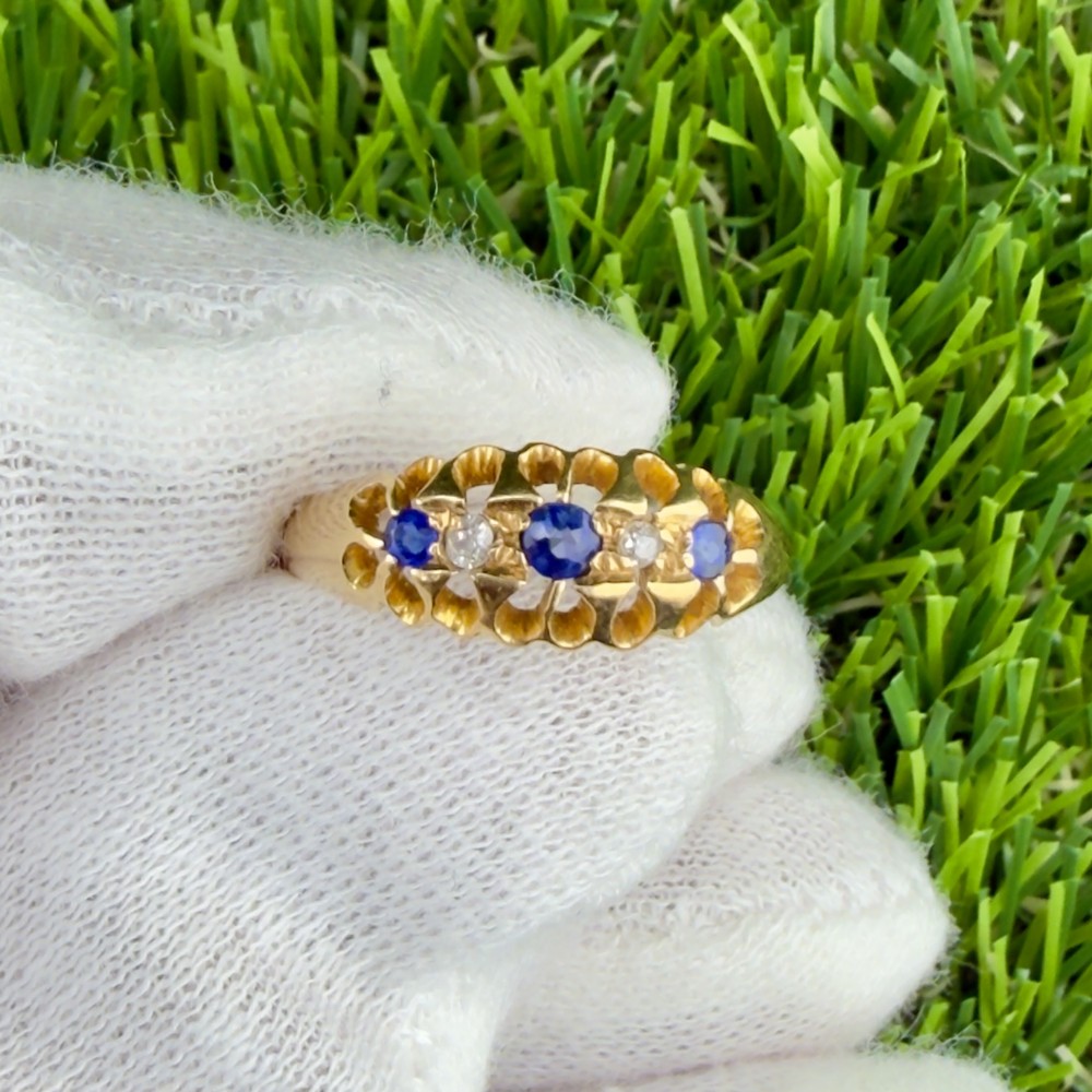 An 18ct yellow gold ring set with three blue sapphires and two small diamonds within a carved scrollwork setting.