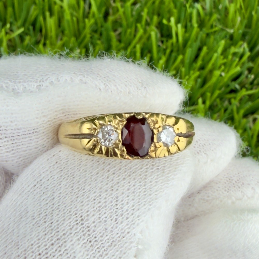 An 18ct yellow gold trilogy ring featuring a central oval dark red garnet flanked by two round diamonds in carved star settings on a wide band.