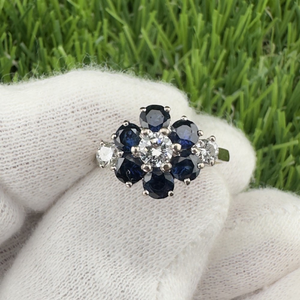 An 18ct white gold cluster ring featuring a central round diamond surrounded by six round dark blue sapphires in a flower motif with diamond accents on the shoulders.