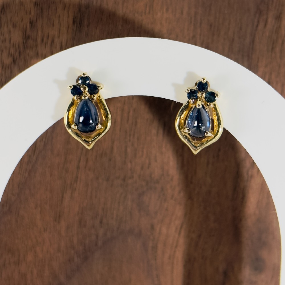 A pair of 9ct yellow gold stud earrings featuring pear-cut cabochon blue sapphires set within textured frames and topped with smaller sapphire accents.