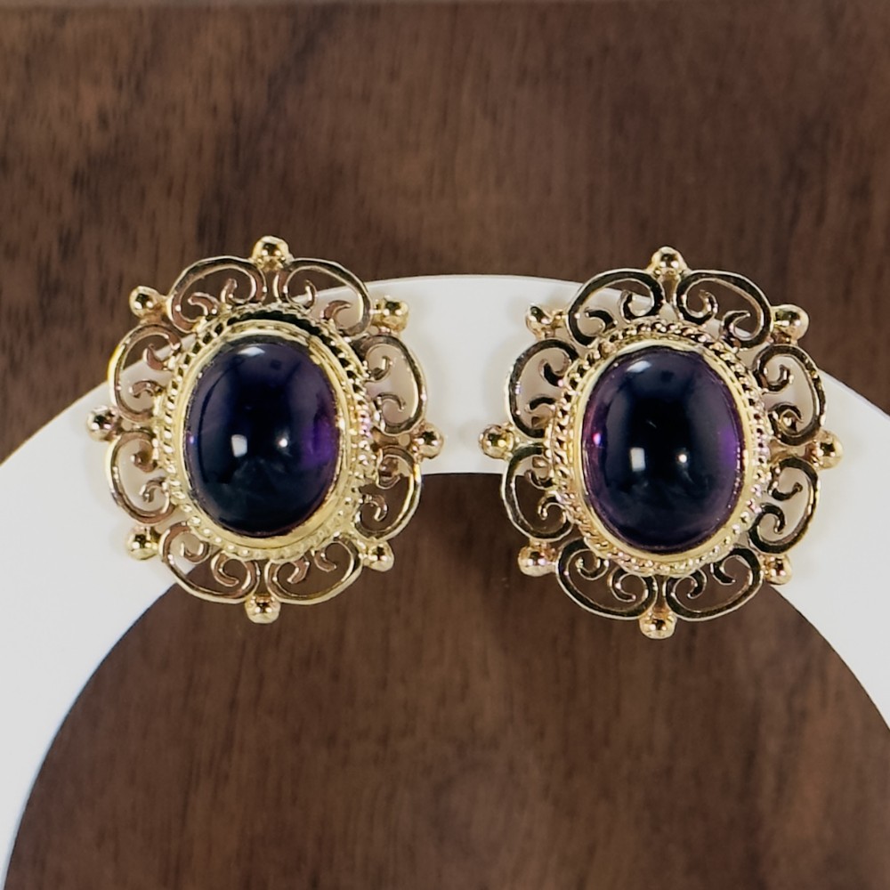 A pair of 9ct gold Edwardian stud earrings featuring central oval cabochon amethysts framed by ornate openwork scrollwork borders.
